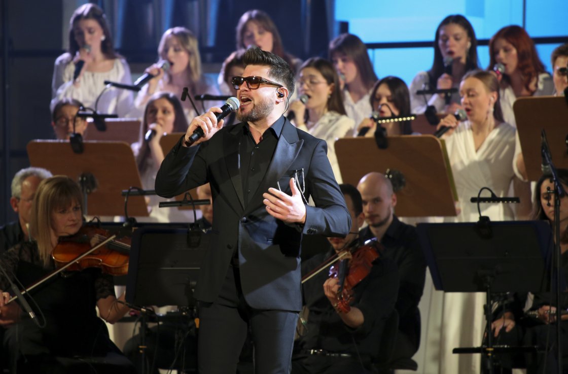 Mężczyzna w eleganckim, czarnym garniturze śpiewa na scenie, trzymając mikrofon. W tle znajduje się chór i orkiestra z muzykami grającymi na skrzypcach. Scena oświetlona jest światłem studyjnym.