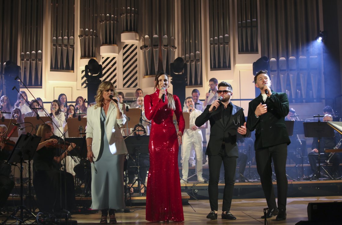 Koncert na scenie z udziałem czterech solistów, orkiestry i chóru. Scena oświetlona kolorowymi światłami, a w tle ogromne organy kościelne. Wokalistka w czerwonej sukni przyciąga wzrok swoim strojem, reszta artystów ubrana elegancko w ciemne i jasne kolor
