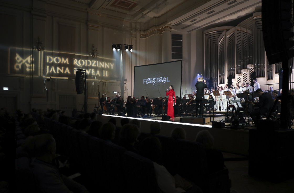 Sala koncertowa wypełniona publicznością. Na scenie występuje orkiestra i chór, obok nich stoi solistka w czerwonej sukni z mikrofonem. Na ścianie wyświetlone światłem logo „Dom Rodzinny Jana Pawła II”. Po prawej stronie duży ekran z napisem „Cały Twój” i