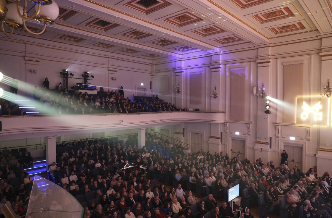 Widok na publiczność w Filharmonii Krakowskiej – tłum ludzi na parterze i balkonie wstaje z miejsc, bijąc brawo w geście owacji na stojąco. Sala wypełniona po brzegi, oświetlona ciepłym światłem sceny. W tle widać klasyczne wnętrza z ozdobnym sufitem i ko