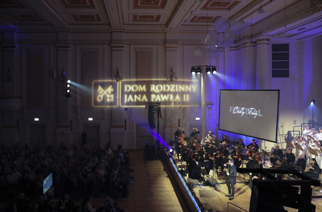 Sala koncertowa wypełniona publicznością. Na scenie występuje orkiestra i chór, obok nich stoi solista z mikrofonem. Na ścianie wyświetlone światłem logo „Dom Rodzinny Jana Pawła II”. Po prawej stronie duży ekran z napisem „Cały Twój” i logotypem festiwal