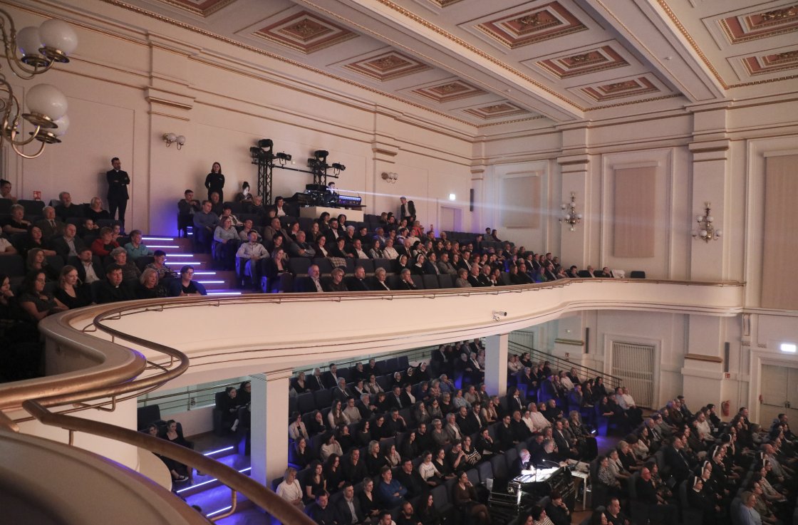 Widok na publiczność zgromadzoną w eleganckiej sali koncertowej z balkonem. Sala wypełniona widzami, scena oświetlona reflektorami, a na ścianie widoczne projekcje świetlne.