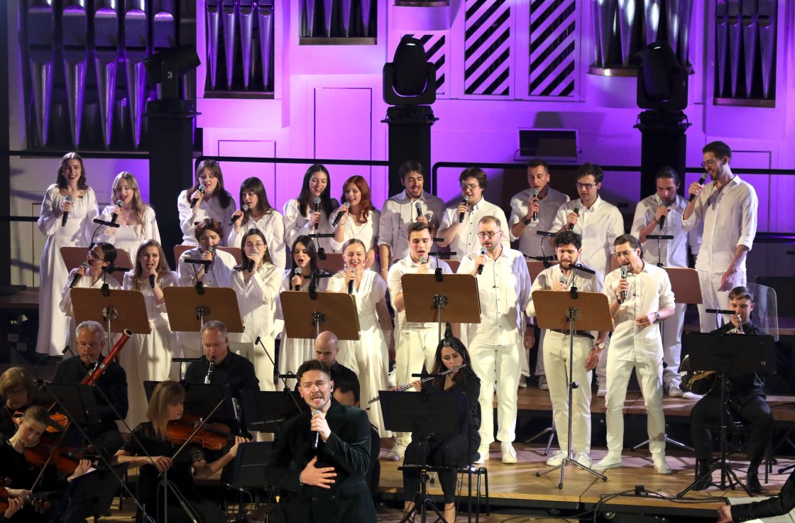 Chór Camerata Jagellonica ubrany na biało. Kobiety i miężczyźni stoją z tyłu sceny. W tle miękkie, fioletowe światło.
