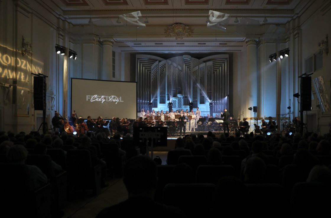 Widok na scenę w eleganckiej sali koncertowej. Sala wypełniona widzami, scena oświetlona reflektorami, a na ścianie widoczne projekcje świetlne.