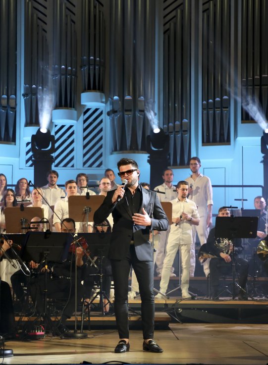 Mężczyzna w eleganckim, czarnym garniturze śpiewa na scenie, trzymając mikrofon. W tle znajduje się chór i orkiestra z muzykami grającymi na skrzypcach. Scena oświetlona jest światłem studyjnym