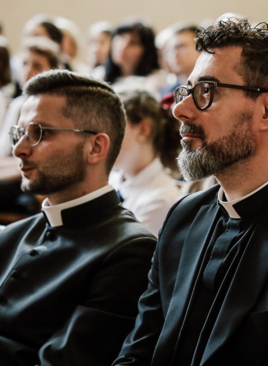 Publiczność siedząca w ławkach podczas finału konkursu. Na pierwszym planie widać dwóch księży w sutannach oraz siostrę zakonną, wszyscy skupieni, słuchają wydarzenia. W tle siedzą uczniowie i dorośli goście.