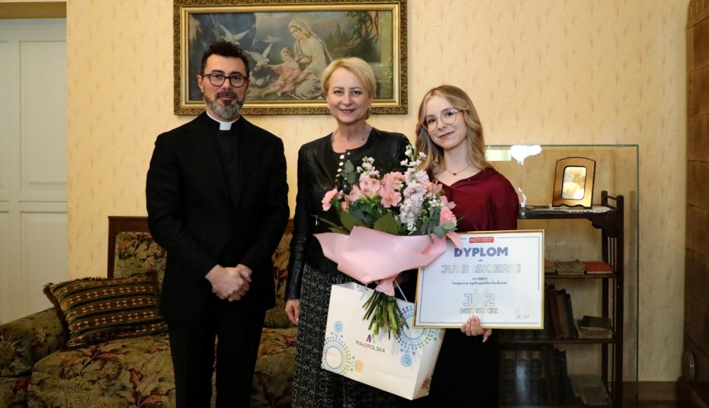 "Trzy osoby stoją obok siebie we wnętrzu stylowo urządzonego pokoju. Po lewej stronie stoi mężczyzna w czarnym garniturze – to ks. dr Łukasz Piórkowski. W środku stoi kobieta w czarnej kurtce i spódnicy – Iwona Gibas, członek Zarządu Województwa Małopolskiego. Po prawej stronie stoi młoda kobieta z długimi blond włosami i okularami – Julia Iskierka, trzymająca bukiet kwiatów, papierową torbę z logotypem Małopolski oraz dyplom za zdobycie pierwszego miejsca w ogólnopolskim konkursie 'JP2inspiruje'. W tle obraz przedstawiający Matkę Boską z Dzieciątkiem oraz drewniane meble.