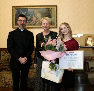 "Trzy osoby stoją obok siebie we wnętrzu stylowo urządzonego pokoju. Po lewej stronie stoi mężczyzna w czarnym garniturze – to ks. dr Łukasz Piórkowski. W środku stoi kobieta w czarnej kurtce i spódnicy – Iwona Gibas, członek Zarządu Województwa Małopolskiego. Po prawej stronie stoi młoda kobieta z długimi blond włosami i okularami – Julia Iskierka, trzymająca bukiet kwiatów, papierową torbę z logotypem Małopolski oraz dyplom za zdobycie pierwszego miejsca w ogólnopolskim konkursie 'JP2inspiruje'. W tle obraz przedstawiający Matkę Boską z Dzieciątkiem oraz drewniane meble.