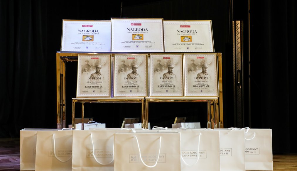 Zbliżenie na zestaw nagród konkursowych ustawionych na eleganckim stoliku. Widać dyplomy z nazwiskami laureatów oraz nagrody w postaci bonów o wartości 2000 zł. Przed stołem ustawiono rząd białych toreb z logotypem „Dom Rodzinny Jana Pawła II”.