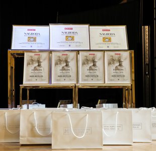 Zbliżenie na zestaw nagród konkursowych ustawionych na eleganckim stoliku. Widać dyplomy z nazwiskami laureatów oraz nagrody w postaci bonów o wartości 2000 zł. Przed stołem ustawiono rząd białych toreb z logotypem „Dom Rodzinny Jana Pawła II”.