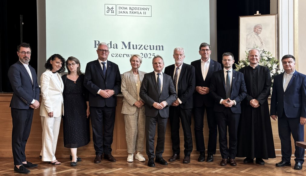 Grupowe zdjęcie wykonane podczas II posiedzenia Rady Muzeum przy Muzeum Dom Rodzinny Ojca Świętego Jana Pawła II w Wadowicach, które odbyło się 17 czerwca 2025 roku. Na pierwszym planie widać jedenastu uczestników spotkania – kobiety i mężczyzn ubranych w eleganckie stroje formalne – stojących na parkiecie w sali z dużym ekranem w tle. Na ekranie widnieje logo Muzeum i napis: „Rada Muzeum, 17 czerwca 2025”. Po prawej stronie ustawiono portret Jana Pawła II i kompozycję kwiatową. Wszyscy pozują do wspólnej fotografii, uśmiechając się do obiektywu.