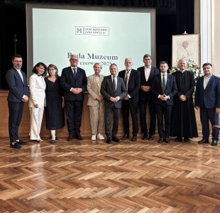 Grupowe zdjęcie wykonane podczas II posiedzenia Rady Muzeum przy Muzeum Dom Rodzinny Ojca Świętego Jana Pawła II w Wadowicach, które odbyło się 17 czerwca 2025 roku. Na pierwszym planie widać jedenastu uczestników spotkania – kobiety i mężczyzn ubranych w eleganckie stroje formalne – stojących na parkiecie w sali z dużym ekranem w tle. Na ekranie widnieje logo Muzeum i napis: „Rada Muzeum, 17 czerwca 2025”. Po prawej stronie ustawiono portret Jana Pawła II i kompozycję kwiatową. Wszyscy pozują do wspólnej fotografii, uśmiechając się do obiektywu.