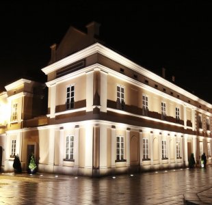 Nocne zdjęcie oświetlonego budynku Muzeum Dom Rodzinny Ojca Świętego Jana Pawła II w Wadowicach. Dwupiętrowy budynek w klasycznym stylu z jasną elewacją, wieloma oknami i ozdobnym podświetleniem. Ulica i plac wokół są mokre po deszczu, odbijają światła. W tle widoczne są inne historyczne budynki centrum miasta.